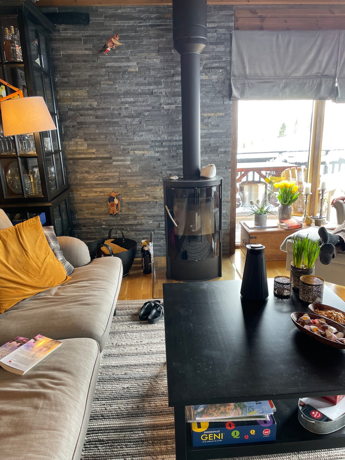 A cozy living area features a modern stove with a sleek black finish, surrounded by textured stone walls. A plush couch adorned with yellow and gray pillows invites relaxation, while a dark coffee table holds decorative items and snacks. Natural light filters through the window treatments.