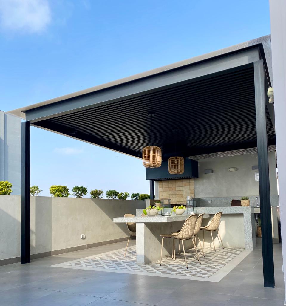 A modern rooftop terrace features a shaded dining area with a large stone table surrounded by comfortable wicker chairs. Two pendant lights hang above, and greenery can be seen along the edges of the terrace, providing a serene outdoor setting.