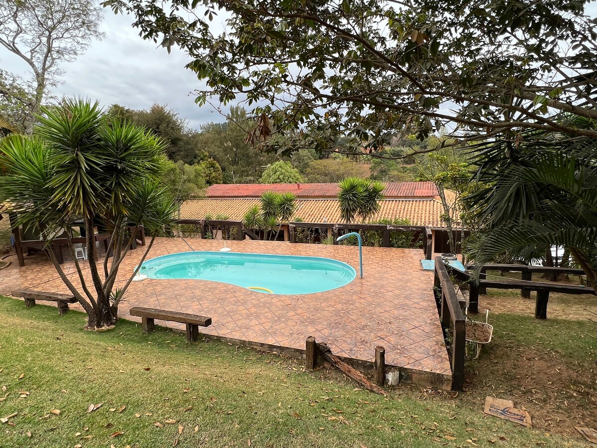 A well-maintained swimming pool is surrounded by lush greenery and palm trees. The pool area features a tiled deck with several benches for seating. A rustic roof can be seen in the background, complemented by a cloudy sky overhead.
