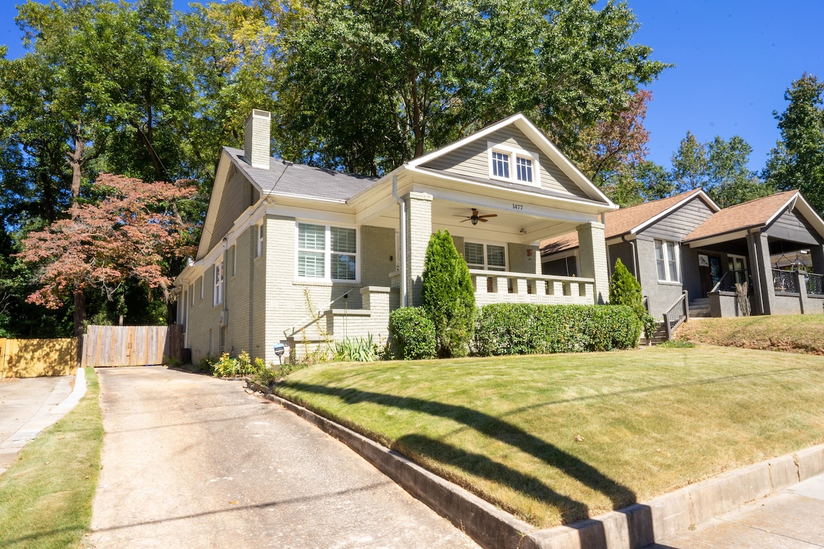 A Craftsman style home is featured with a light-colored exterior and a welcoming front porch. The well-maintained lawn includes trimmed hedges and small landscaping elements, while trees provide shade to the entrance. The nearby street indicates a residential neighborhood.
