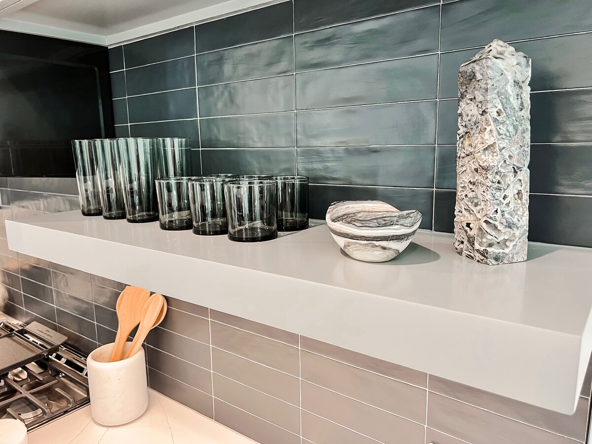 A sleek kitchen counter displays an arrangement of tinted glass drinking glasses alongside unique decorative items. A smooth bowl with waves of grey and white sits next to a textured stone piece, all set against a contemporary dark tiled backsplash.