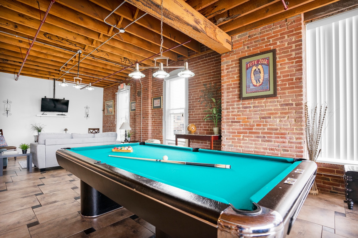 A vibrant pool table with a turquoise surface occupies the center of the open-concept space, surrounded by exposed brick walls and wooden beams. Wall art and a large TV are visible in the background, contributing to a relaxed atmosphere.