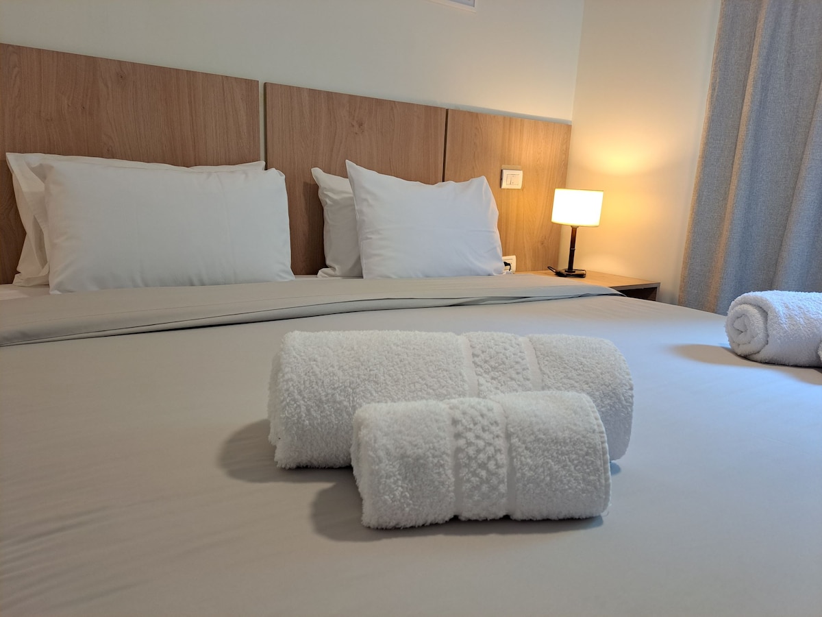 A neatly made bed is presented with two white towels folded neatly at the foot. The headboard features a natural wood finish, and a soft bedside lamp emits warm light in the background, complemented by a light-colored curtain that softly frames the window.