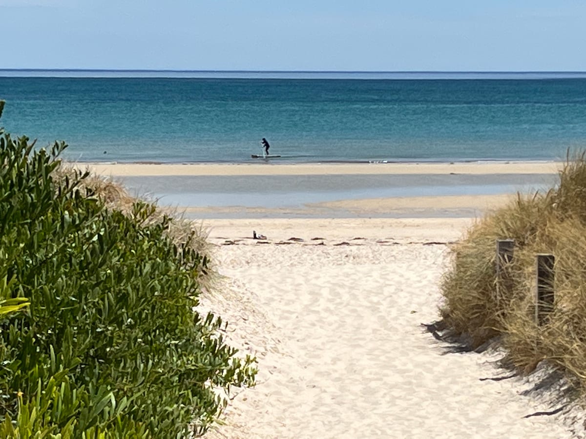 A sandy pathway leads to a serene beach featuring calm turquoise waters. A lone surfer can be seen riding gentle waves in the distance, while lush greenery frames the scene, creating a peaceful coastal atmosphere.