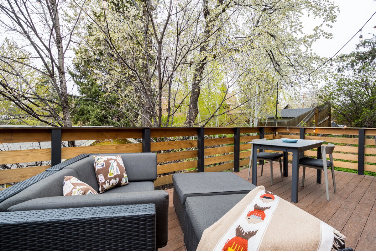 A spacious outdoor deck features a comfortable sofa and a dining table for four. Mature trees surround the space, providing natural privacy. A patterned throw blanket is casually draped over the sofa, complementing the serene outdoor setting.