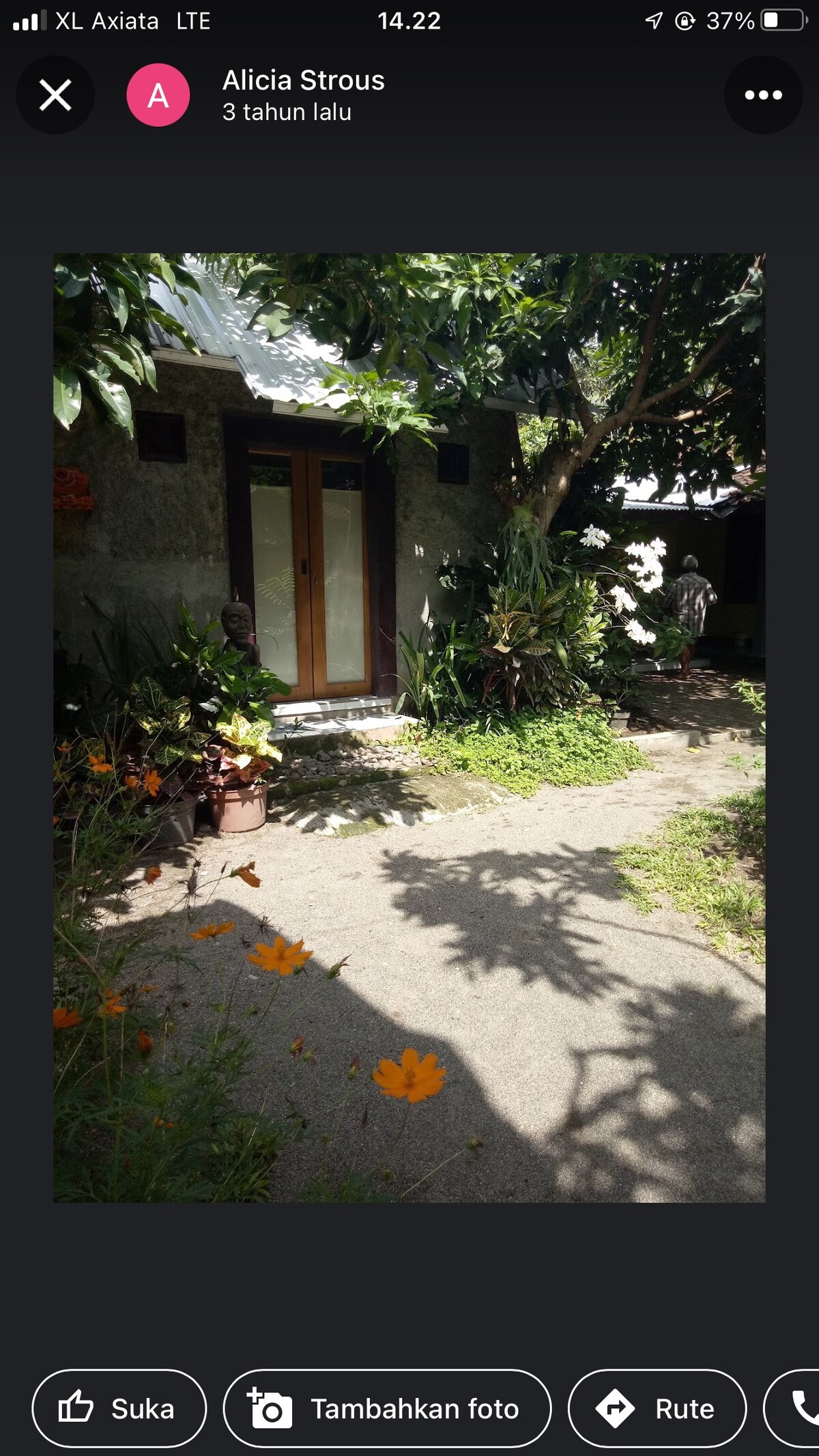A pathway leads to a serene entrance framed by lush greenery and vibrant flowers. Sunlight filters through the leaves, illuminating the area. The entrance features wooden doors that blend harmoniously with the natural surroundings.