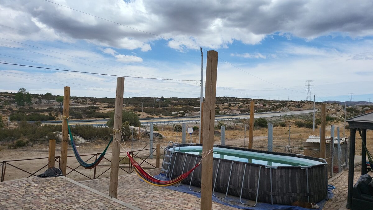 An outdoor area features a circular above-ground pool surrounded by wooden supports. Two colorful hammocks are suspended nearby, providing a relaxed space to enjoy the surrounding landscape. The dry terrain and distant hills are visible under a partly cloudy sky, with power lines running alongside a nearby road.