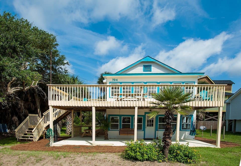 The exterior of a two-story beach house is presented, featuring a spacious deck shaded by a roof. Palm trees and greenery surround the property. A set of stairs leads to the elevated entrance, while a bench is placed on the ground level patio area beneath the deck.