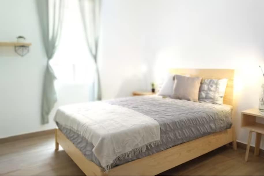 A room features a spacious bed with a light-colored blanket and decorative pillows. Natural light enters through a partially draped window. A wooden nightstand with a small plant is positioned beside the bed, complementing the minimalist decor.