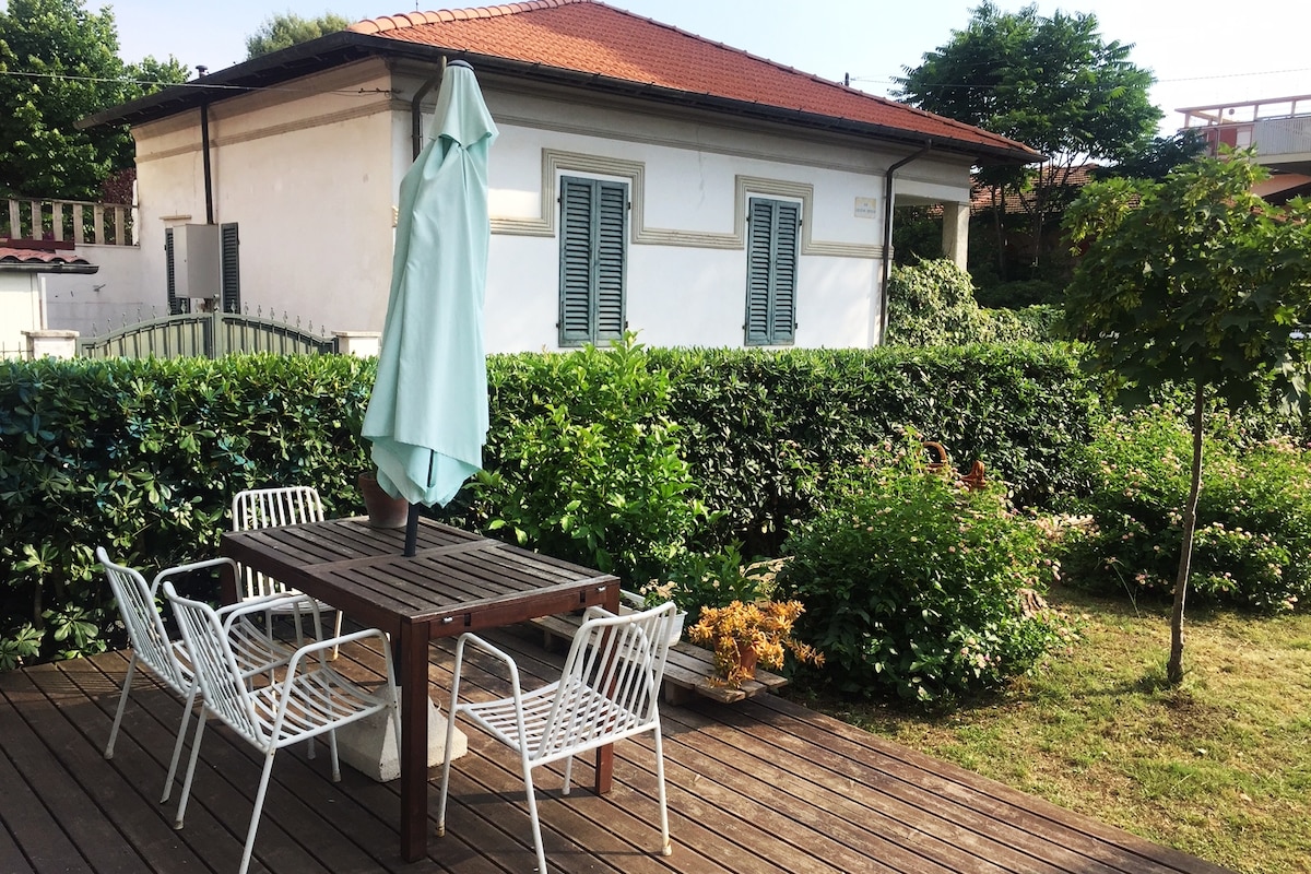 An outdoor dining area features a wooden table surrounded by four metal chairs, sheltered by a light green umbrella. Lush greenery, including hedges and small trees, frames the space, creating a serene environment. The bright exterior of the apartment is visible in the background.