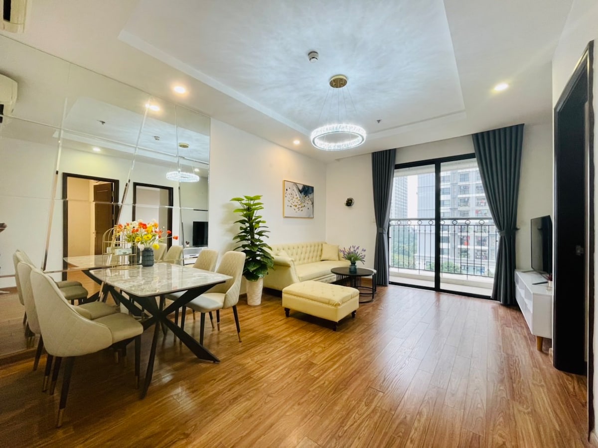 A bright living area features a large window providing natural light. A comfortable sectional sofa and a dining table with chairs are positioned across from a television. Decorative plants add greenery, while a mirrored wall enhances the sense of space.