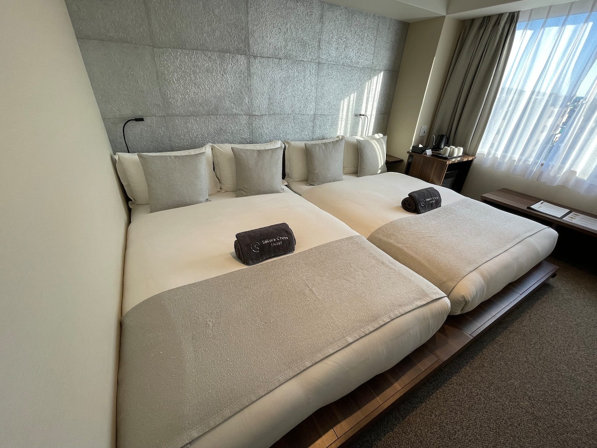 A bright guest room features two spacious beds with neutral-colored bedding and decorative pillows. Soft lighting is provided by the nearby window, dressed with sheer curtains. A minimalistic wooden table is positioned alongside the beds, enhancing the room's contemporary design.