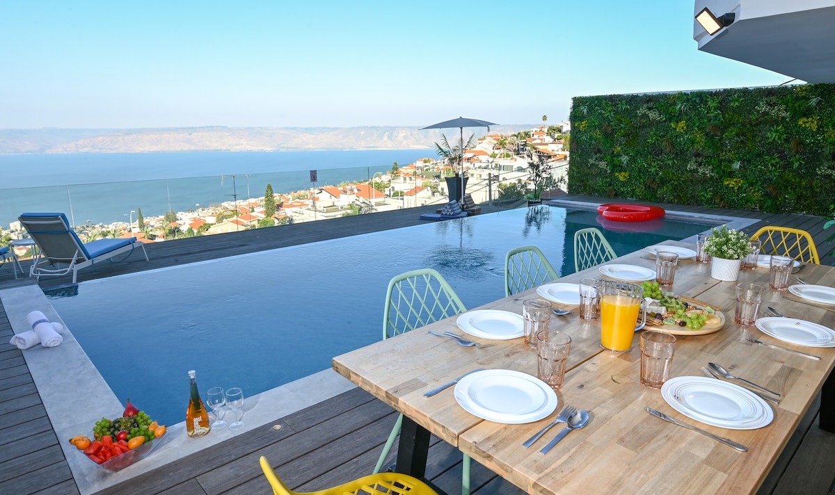 An outdoor dining area is set with plates and glassware around a wooden table. A large infinity pool reflects the surrounding view, including the Sea of Galilee and distant hills. Colorful chairs and a sun umbrella enhance the inviting atmosphere, with fresh produce visible on the table.