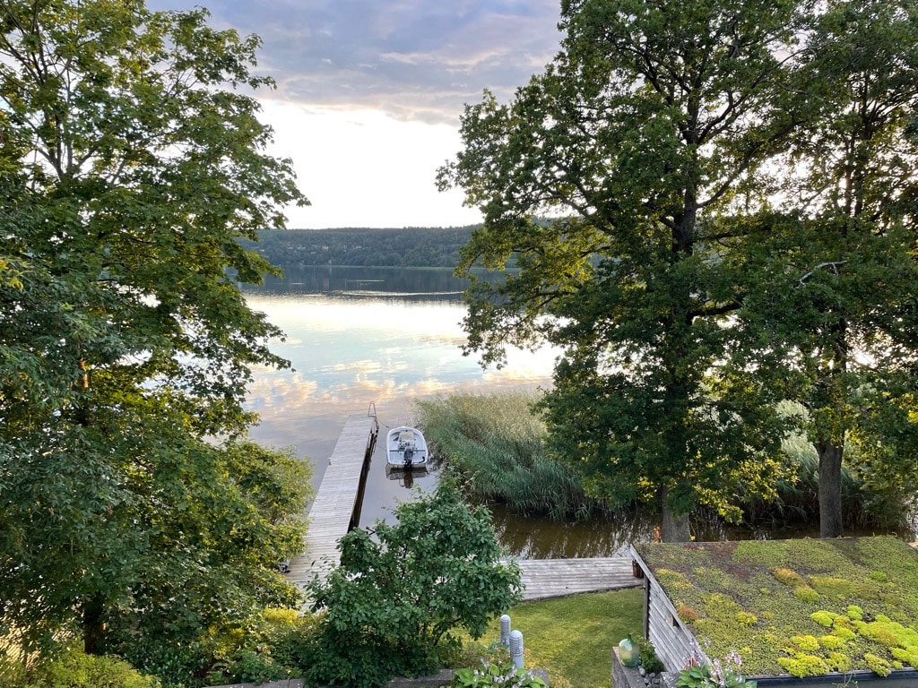 A serene view of a private dock extending into the calm lake, surrounded by lush trees and tall grasses. The sky reflects soft colors as the sun sets, enhancing the tranquil atmosphere created by the natural surroundings.