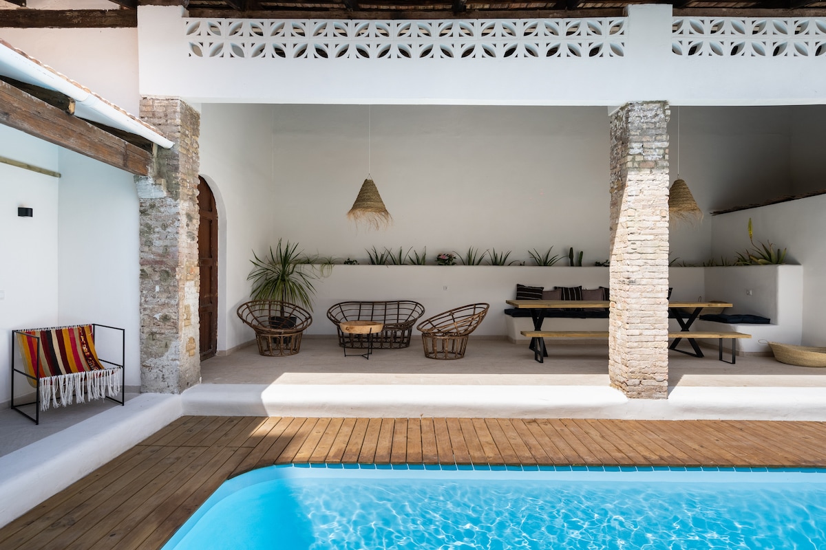 An open-air lounge area features natural materials with rattan chairs and a long wooden dining table. The space is framed by rustic stone columns and layered with greenery, overlooking a private pool that reflects the surrounding light.