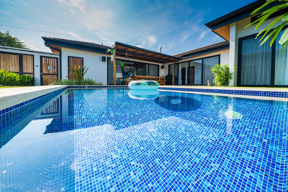 A serene swimming pool is visible, surrounded by a spacious deck and lush greenery. The clear water reflects the sky and features a floating inflatable for relaxation. Large glass doors of the house provide a view into the inviting living area.