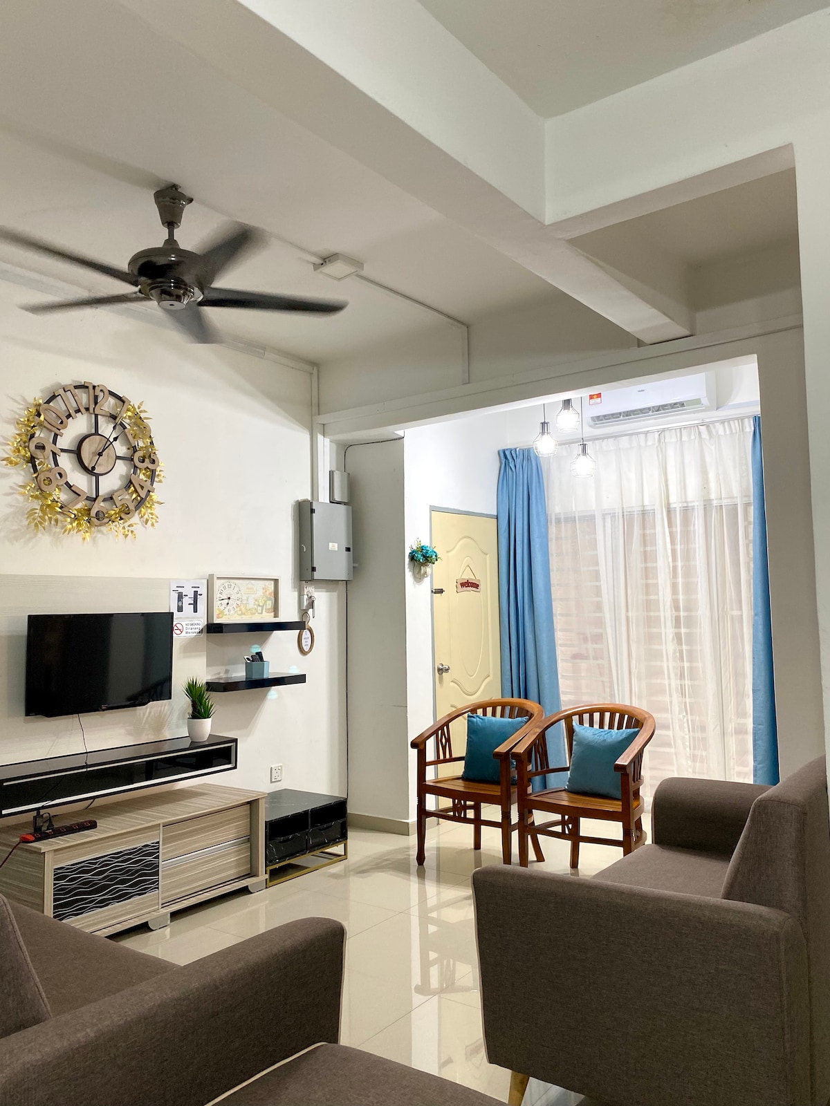 A cozy living area is presented, featuring a wall-mounted television and a decorative clock. Two cane chairs are positioned beside a window with light curtains, allowing natural light to enter. Two gray sofas provide additional seating, enhancing the room's functionality.