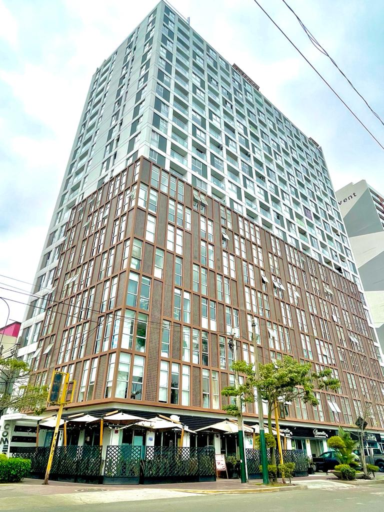 A modern multi-story building features a blend of glass and brick facade, providing a contemporary urban aesthetic. Large windows reflect the nearby surroundings, while trees and greenery line the street level, enhancing the building's appeal and inviting atmosphere.