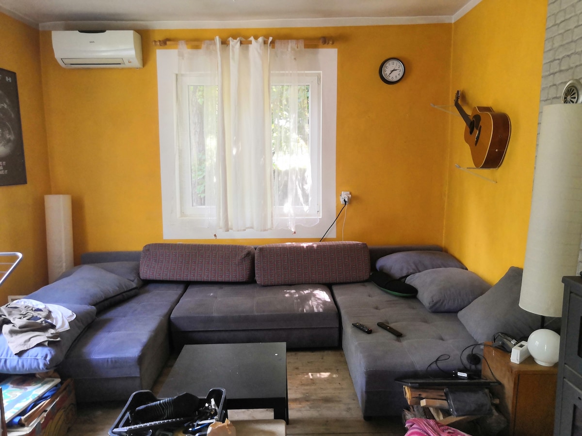 A spacious corner sofa is positioned against a bright yellow wall, providing ample seating. A wooden coffee table sits in front, accompanied by a clock and guitar hanging on the wall. Natural light enters through the window, adorned with light curtains.