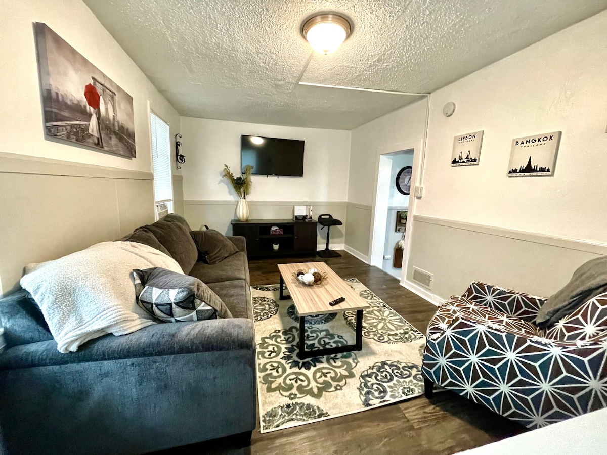 A comfortable living area is presented with a dark gray sofa and a patterned accent chair. A central coffee table holds decorative items, and a wall-mounted television is visible. Light walls and a soft area rug create a welcoming environment.