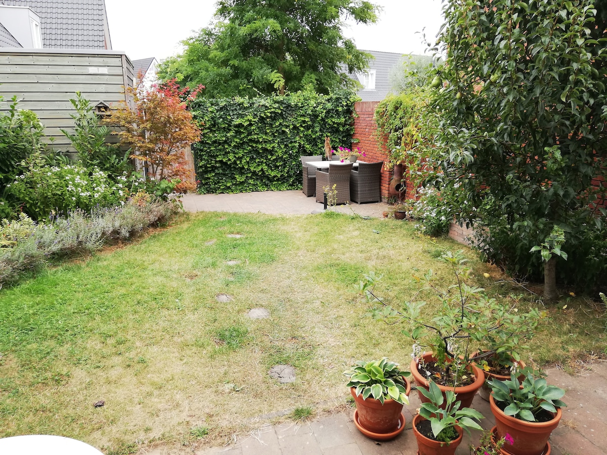 A garden space is depicted, featuring a small seating area with a table and chairs surrounded by greenery. Flowering plants and shrubs are arranged in pots along the edges. A well-maintained lawn extends towards the back, bordered by a mix of foliage and brick walls.