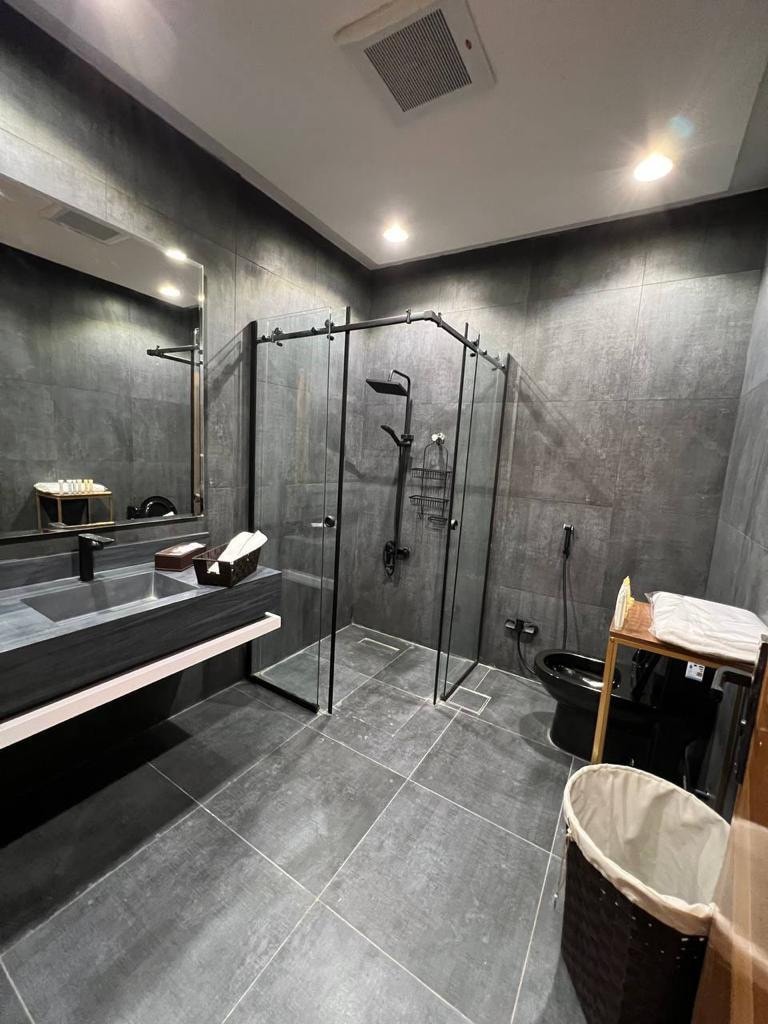A modern bathroom features dark stone walls and sleek finishes. A glass-enclosed shower with black fixtures stands adjacent to a clean countertop with a large mirror. A neutral-colored laundry hamper is positioned nearby, and a stylish storage unit complements the space.