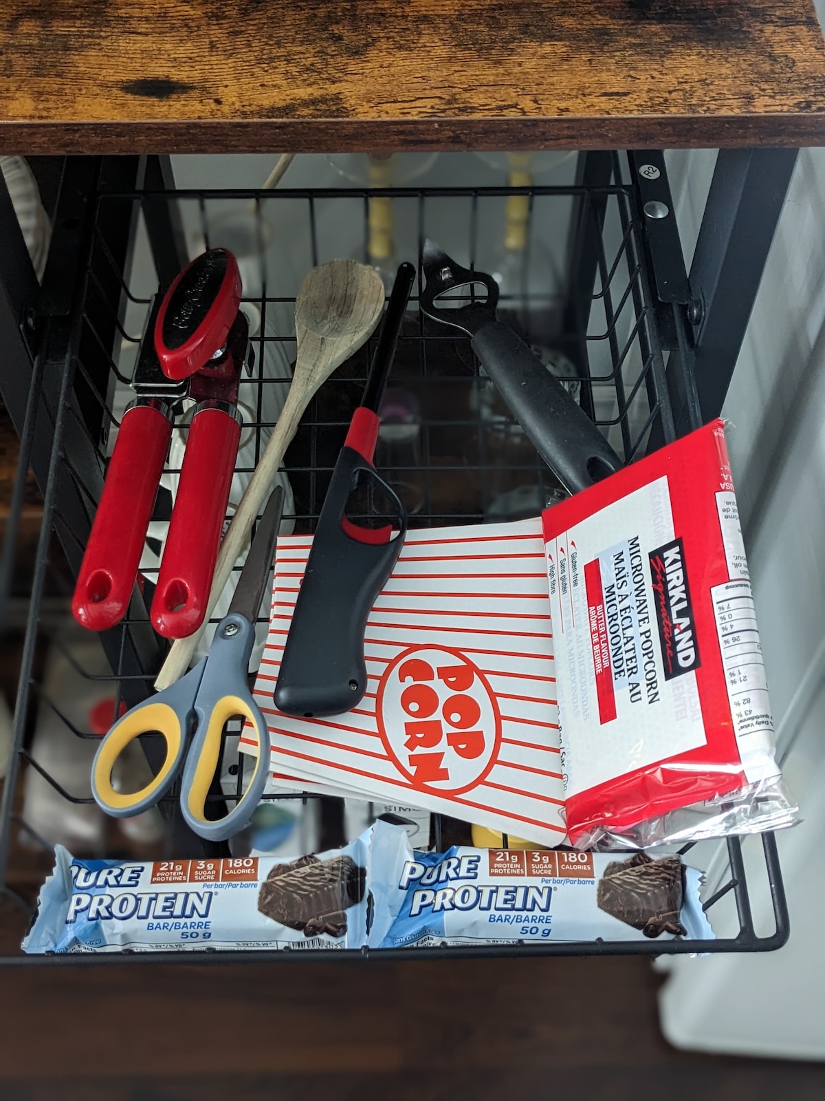 A container holds various kitchen tools and snacks. Visible items include a red handled can opener, a wooden spoon, a pair of scissors, a popcorn box, and two protein bars. A lighter and a bottle opener are also present among the items.