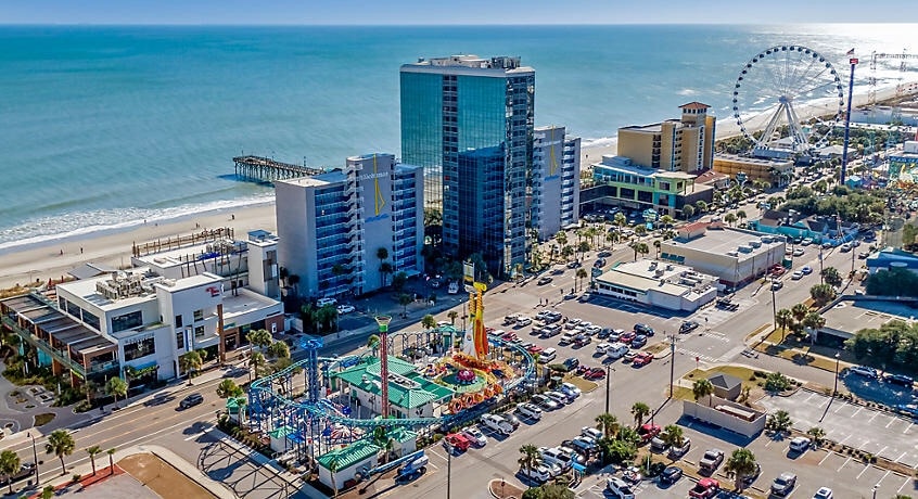 An aerial view captures the vibrant Myrtle Beach area, featuring a colorful amusement park with rides and attractions. Nearby, the ocean sparkles under sunlight, while the beach and pier extend along the coastline. The skyline includes modern buildings, creating a lively atmosphere.