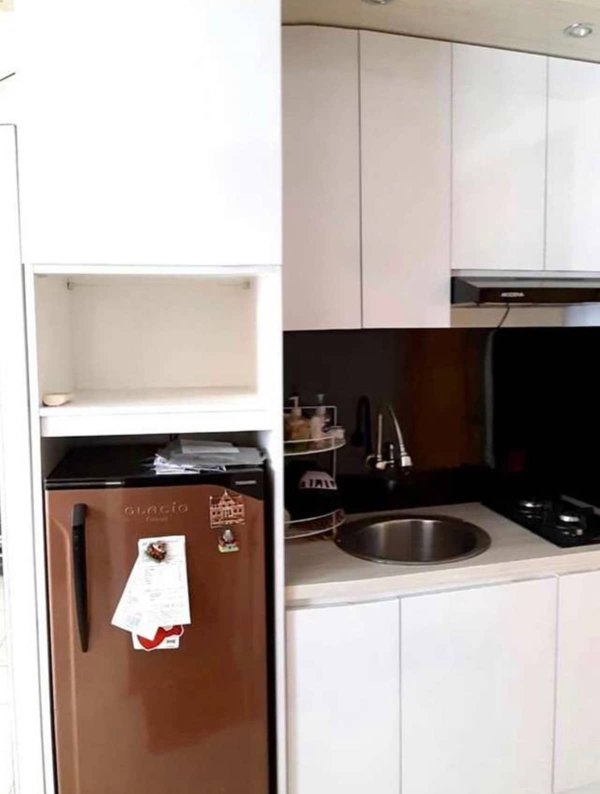 A compact kitchenette is arranged with modern cabinetry and a sleek black backsplash. A stainless steel sink sits beside an under-counter refrigerator. A microwave is positioned above the sink, while various utensils and kitchenware are organized on the shelves.