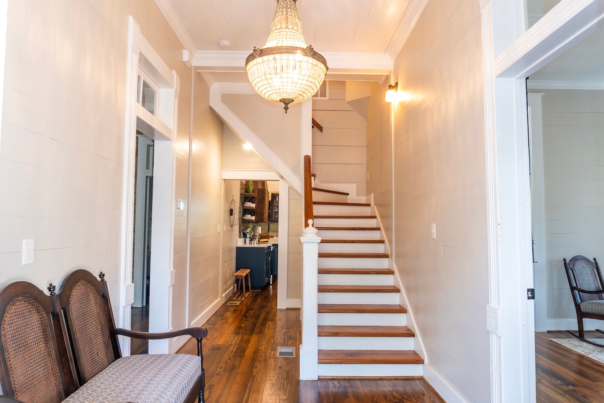 An inviting entryway features a chandelier above a staircase with wooden steps leading to the second floor. Two elegant chairs are positioned near the wall, offering a cozy spot for guests to relax. Natural light illuminates the warm wooden flooring.