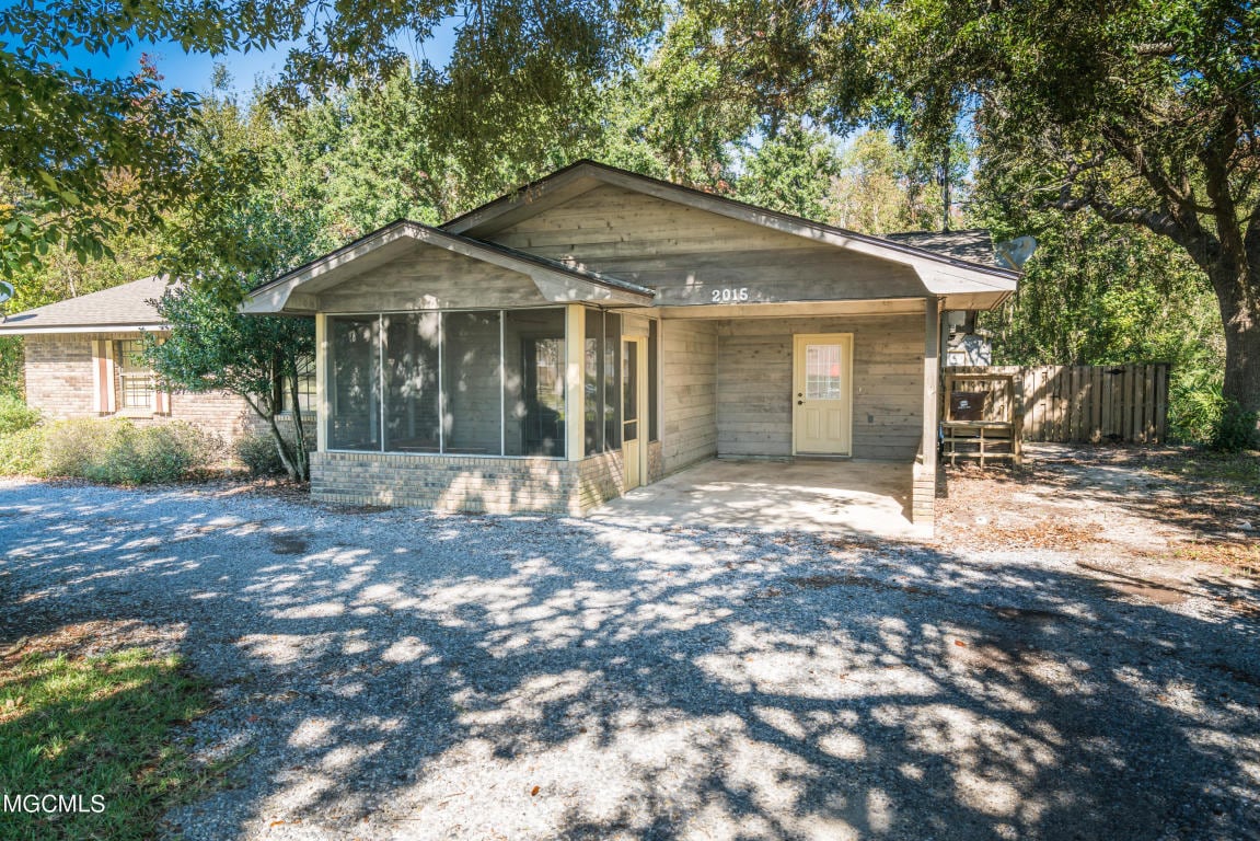 The exterior of a single-story home is presented, featuring a front porch with a screened area. A gravel driveway provides access to the property, surrounded by lush trees that cast dappled shadows on the ground.