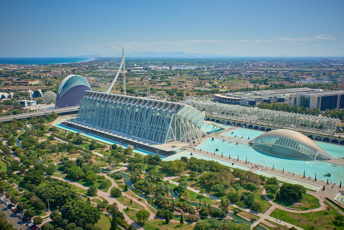 An aerial view captures the striking architecture of the City of Arts and Sciences in Valencia, surrounded by lush greenery and waterways. The complex features futuristic structures, set against a clear blue sky, with the coastline visible in the distance.