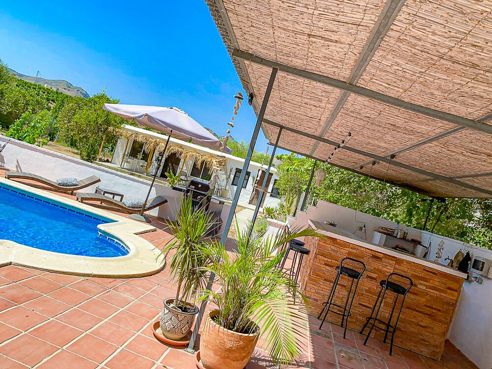 A spacious outdoor area features a private pool surrounded by a sun terrace. A shaded bar area with stools is situated nearby, complemented by vibrant greenery. An umbrella provides shade over a lounge chair, enhancing the relaxing atmosphere near the pool.