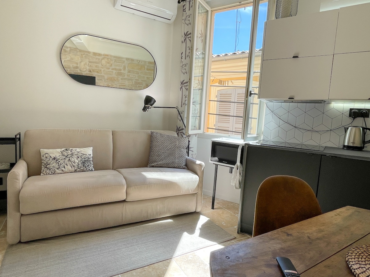 A modern living space features a light-colored sofa with decorative cushions, a sleek dining table, and a fully equipped kitchenette. Natural light fills the room through a large window, revealing a view of traditional architecture. Decorative elements include a round mirror and subtle patterned curtains.