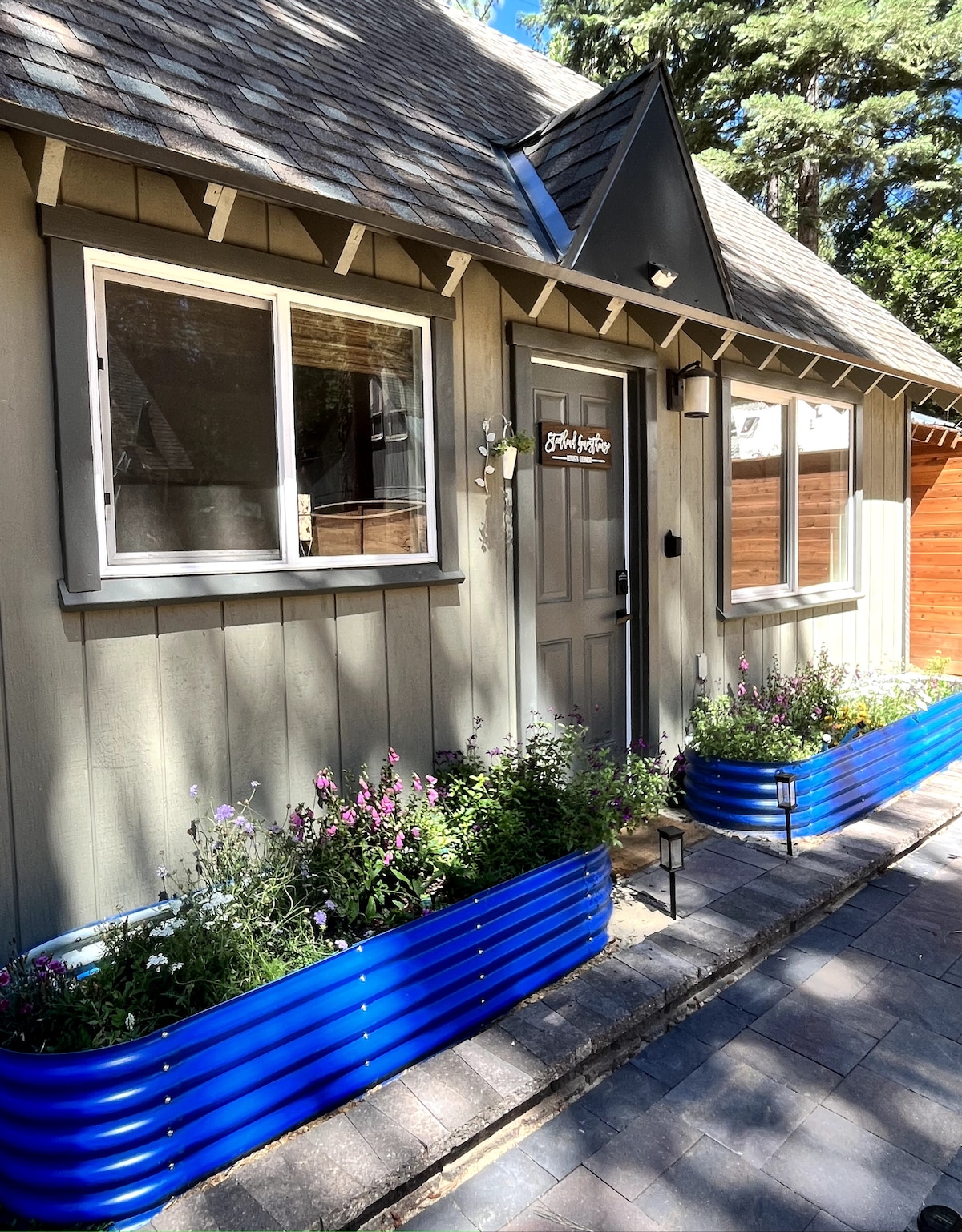 A charming entrance to the Steelhead Guesthouse is framed by vibrant blue planters filled with colorful flowers. Natural stone pavers lead to a welcoming door, accented by a simple light fixture and a wooden sign displaying the property name.