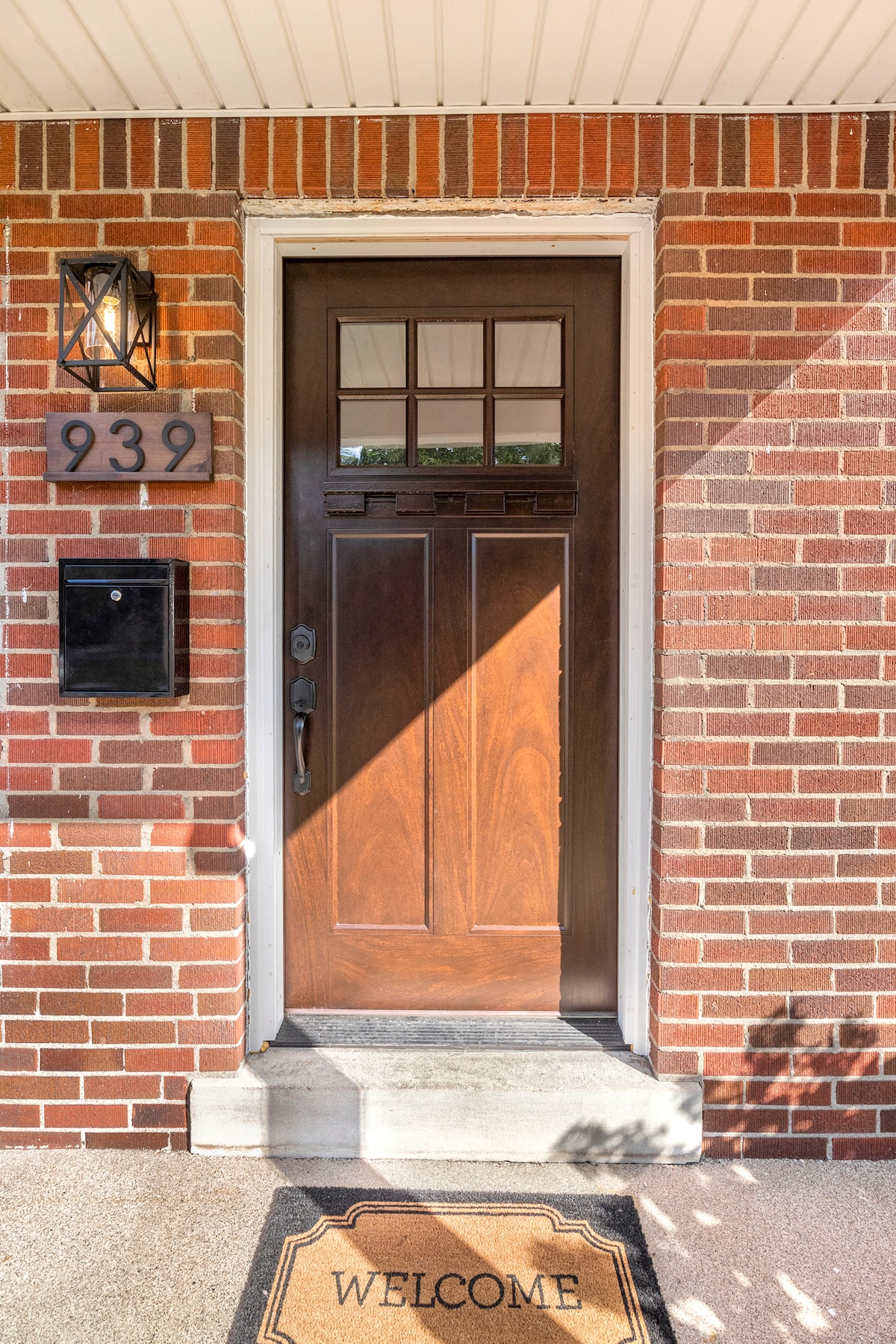 A solid wooden door with a decorative window above it is framed by a brick exterior. The house number is displayed prominently, and a welcoming doormat is placed at the entrance, inviting visitors into the home.