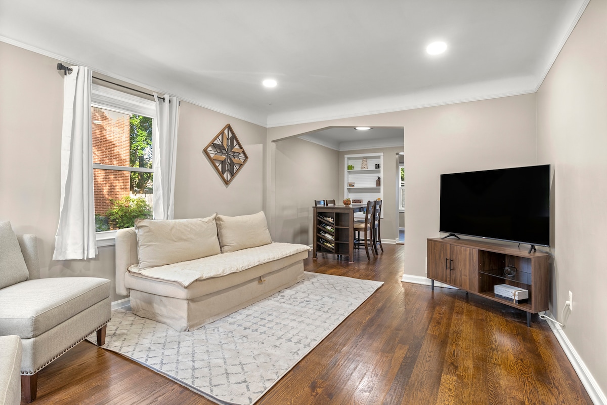 A light-filled living area showcases a neutral sofa and a decorative mirror on the wall. A cozy area rug covers the hardwood floor. To the right, a dining space is visible, featuring a table and stools, complemented by shelves in the background.