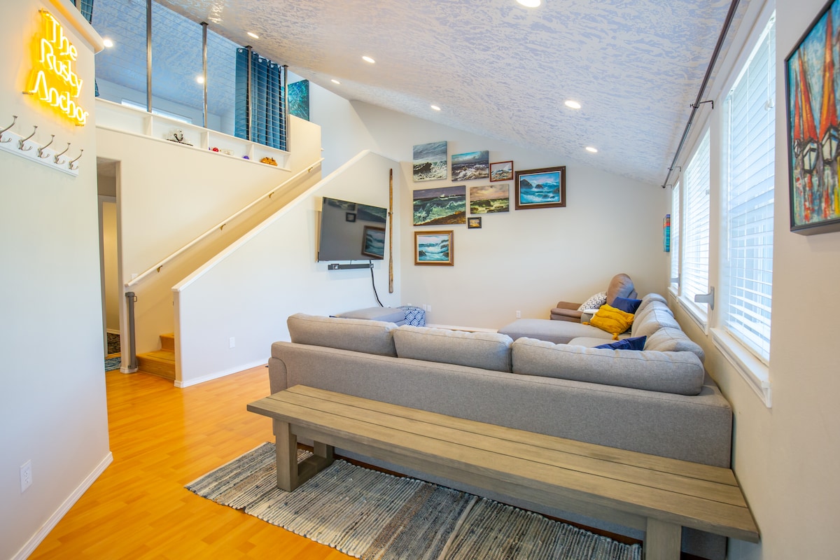 A spacious living area features a large sectional sofa with accent pillows, complemented by a wooden coffee table. Sunlight filters through multiple windows, illuminating artwork displayed on the walls. A staircase is visible in the background, leading to the upper level.