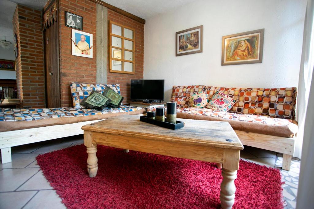 A cozy living area features two wooden couches adorned with colorful cushions, complemented by a rustic wooden coffee table. A television is positioned against a brick wall, while art pieces are displayed on nearby surfaces. A vibrant red rug adds warmth to the space.