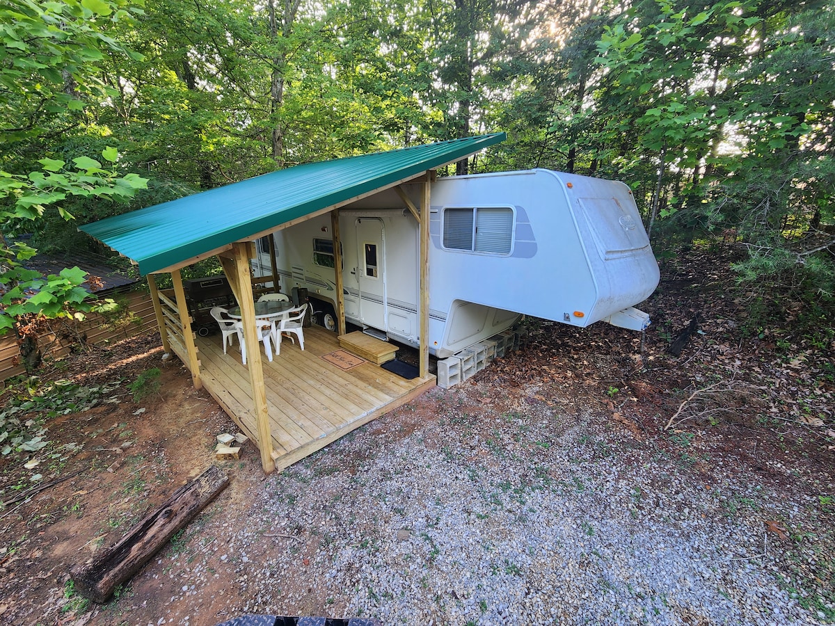 A cozy camper is shown nestled among trees, featuring a covered porch with a wooden deck. A small outdoor seating area with white chairs is visible, along with gravel ground. The camper’s exterior is light-colored with a distinctive green roof.