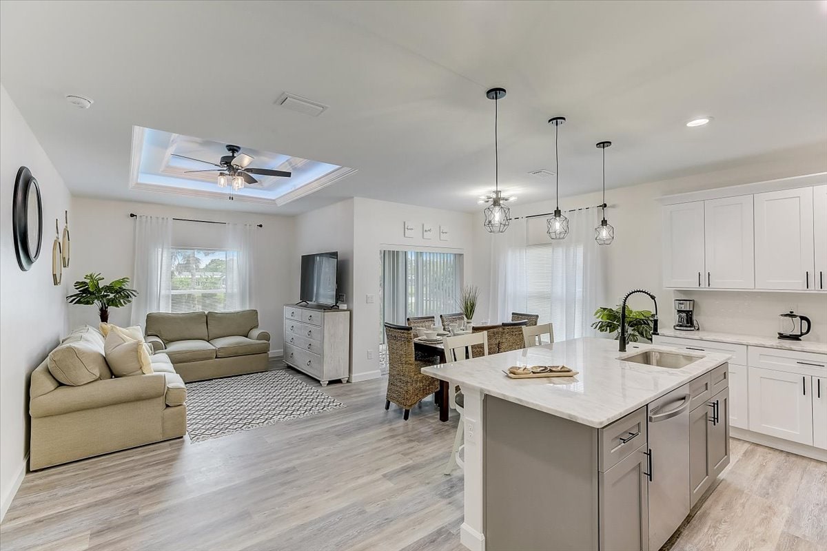 A spacious living area is equipped with soft seating and a large flat-screen television. Natural light enters through multiple windows, illuminating the room. A modern kitchen features an island with seating and is complemented by light cabinetry and a dining space for six.
