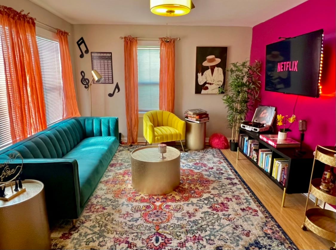 A vibrant living space features a turquoise sofa and a yellow accent chair situated around a round, metallic coffee table. Colorful curtains complement a striking pink accent wall, while musical notes adorn one side. A TV displaying Netflix is mounted above a low media console filled with books.