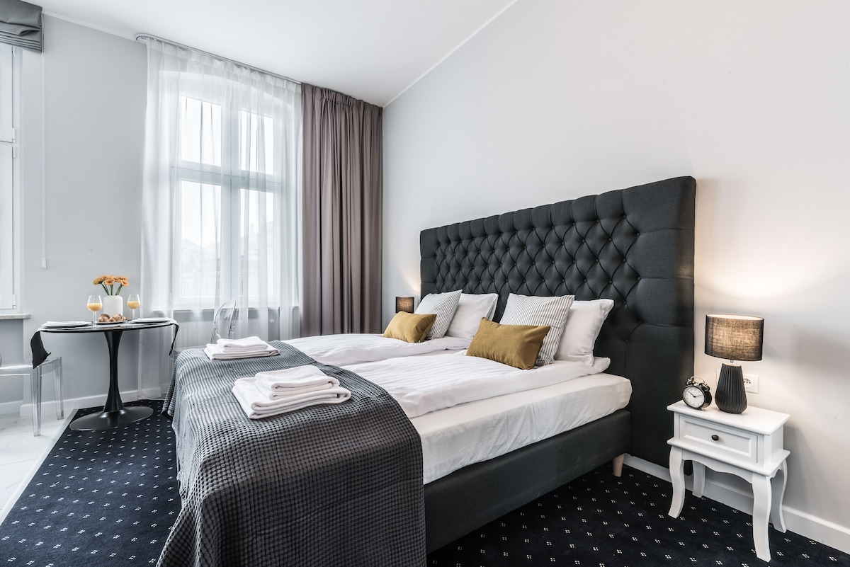 The room features a spacious bed with a tufted, dark headboard, complemented by decorative cushions. A side table with a lamp and an alarm clock is positioned next to the bed. Natural light fills the space through large windows dressed in sheer curtains, while a small table is set for two.