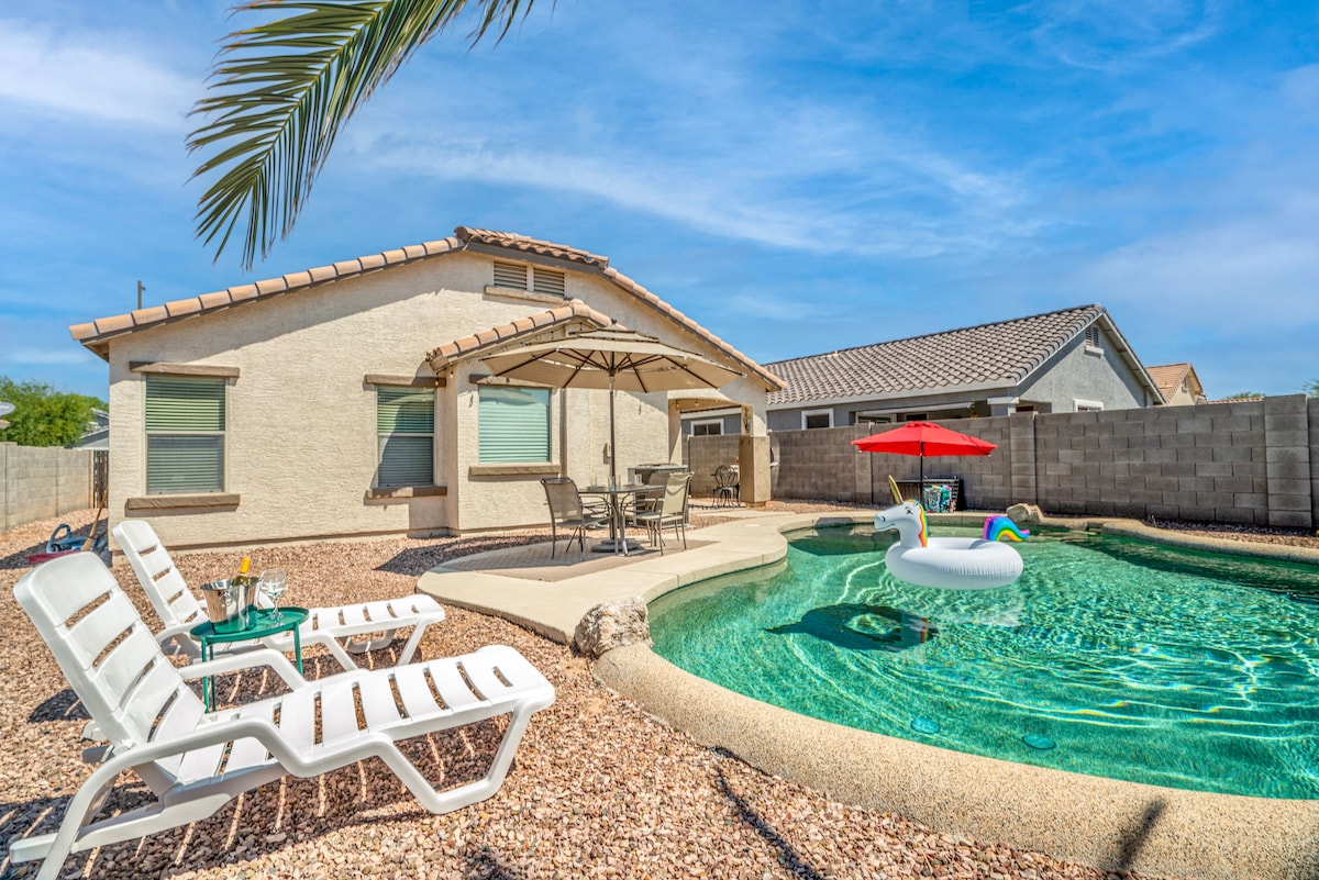 A private pool features clear water surrounded by a pebbled patio with lounging chairs. An umbrella-shaded table and chairs are present nearby. A colorful inflatable unicorn floats in the pool, with the home's exterior visible in the background.