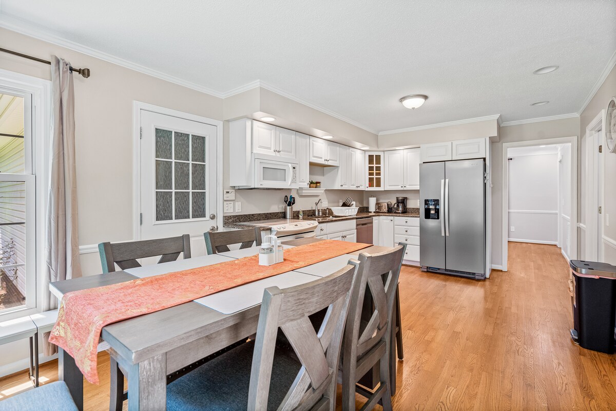 A modern kitchen features a spacious layout with white cabinetry and stainless-steel appliances. A large dining table is set with a centerpiece, surrounded by wooden chairs. Natural light enters through multiple windows, enhancing the inviting atmosphere of the space.