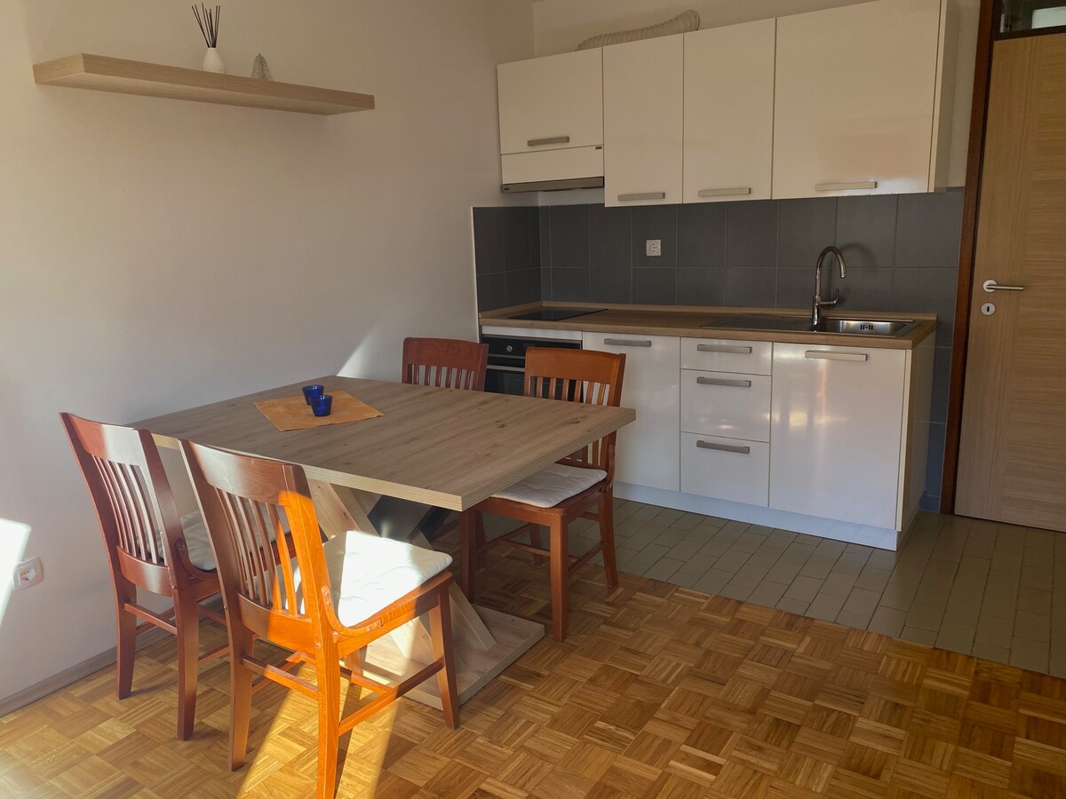A modern kitchen area features a wooden dining table with four matching chairs. The kitchen cabinetry is in a clean white finish, complemented by a contrasting dark countertop and backsplash. Natural light fills the space, highlighting the warm tones of the parquet flooring.