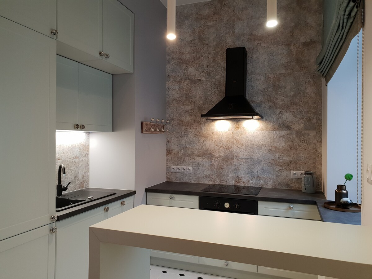 A modern kitchen features sleek white cabinetry and a contrasting dark countertop. A black hood vent is mounted above the stovetop, with two pendant lights illuminating the workspace. The kitchen is completed with a black sink and a subtle textured wall in neutral tones.