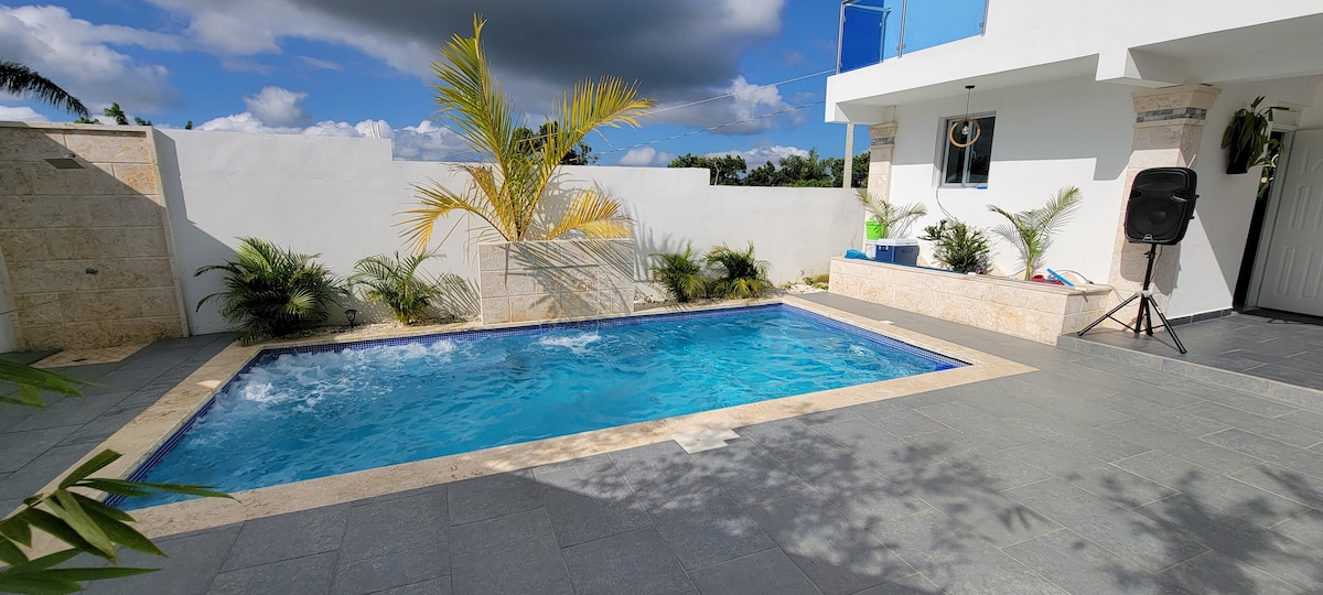 A private pool is surrounded by tile flooring and tropical plants. Clear blue water glimmers under sunlight. A portion of the pool area is shaded by a palm tree. The backdrop features white walls and a nearby outdoor space, enhancing the sense of openness.