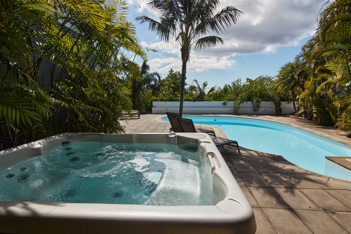 A smooth surface Jacuzzi is positioned beside an inviting swimming pool, surrounded by lush greenery and palm trees. The spacious patio area features lounge chairs, and ample natural light is present, with clouds partially visible in a blue sky.