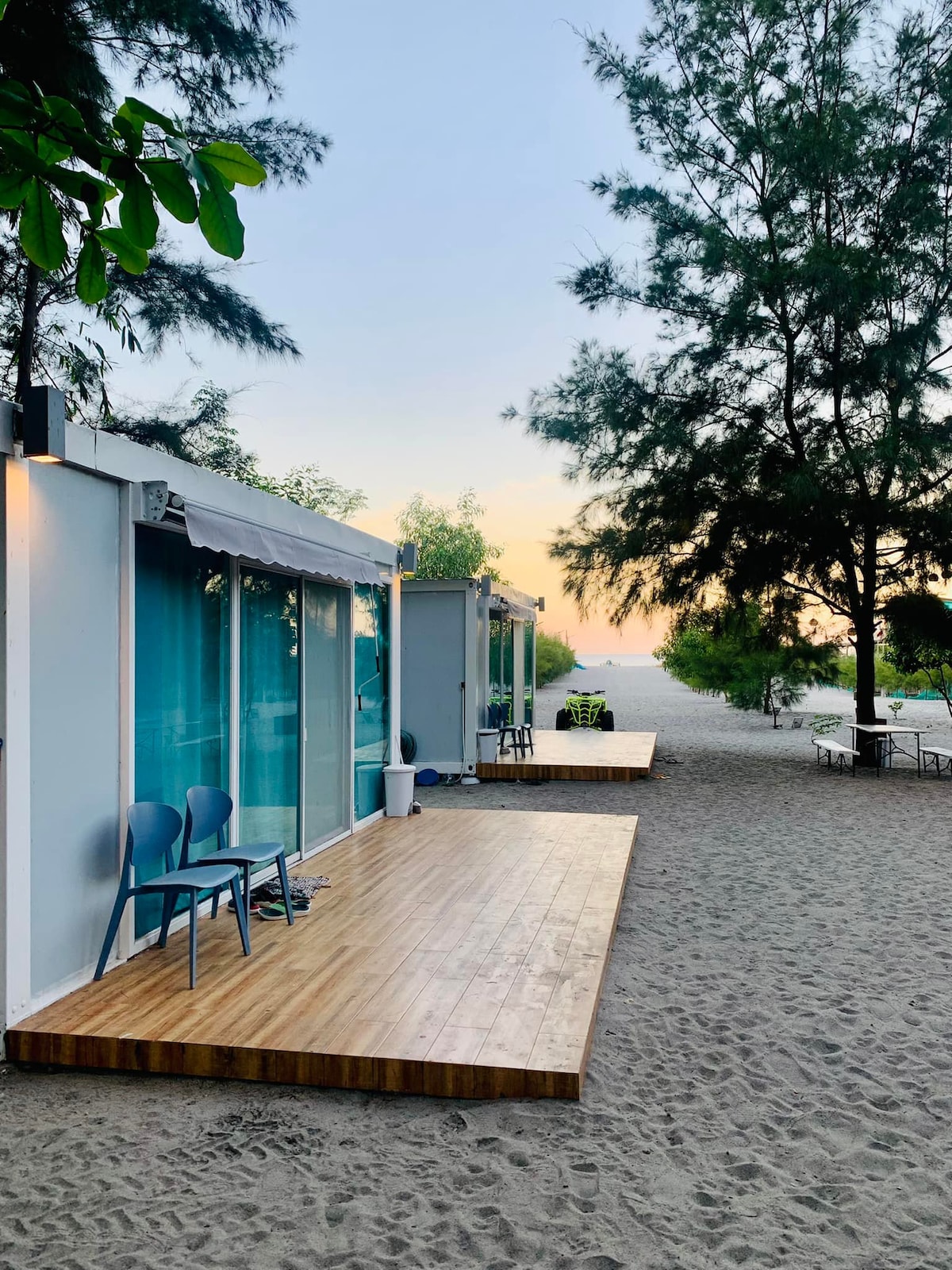 Three modern cubes are positioned on a sandy area, each with wooden decks and large glass doors, reflecting the surroundings. Blue chairs are arranged on the decks, with greenery and trees in the background, enhancing the coastal ambiance as the sun sets.
